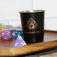 Speakeasy Dice Metal Shot Glass with a matte black finish, shown next to a d4 die for scale. The gold Speakeasy Dice logo stands out, making this durable shot glass perfect for Dungeons & Dragons game nights or barware collections.