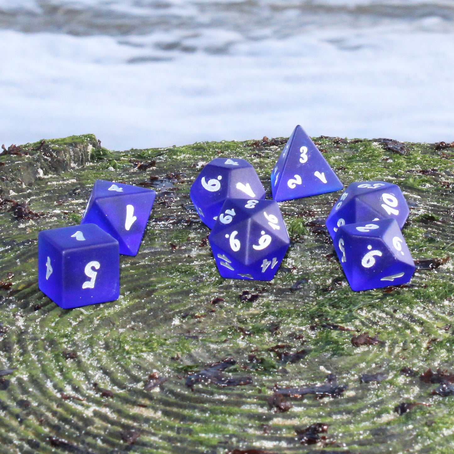 A set of dark blue translucent glass RPG dice with silver numbering on a grassy surface