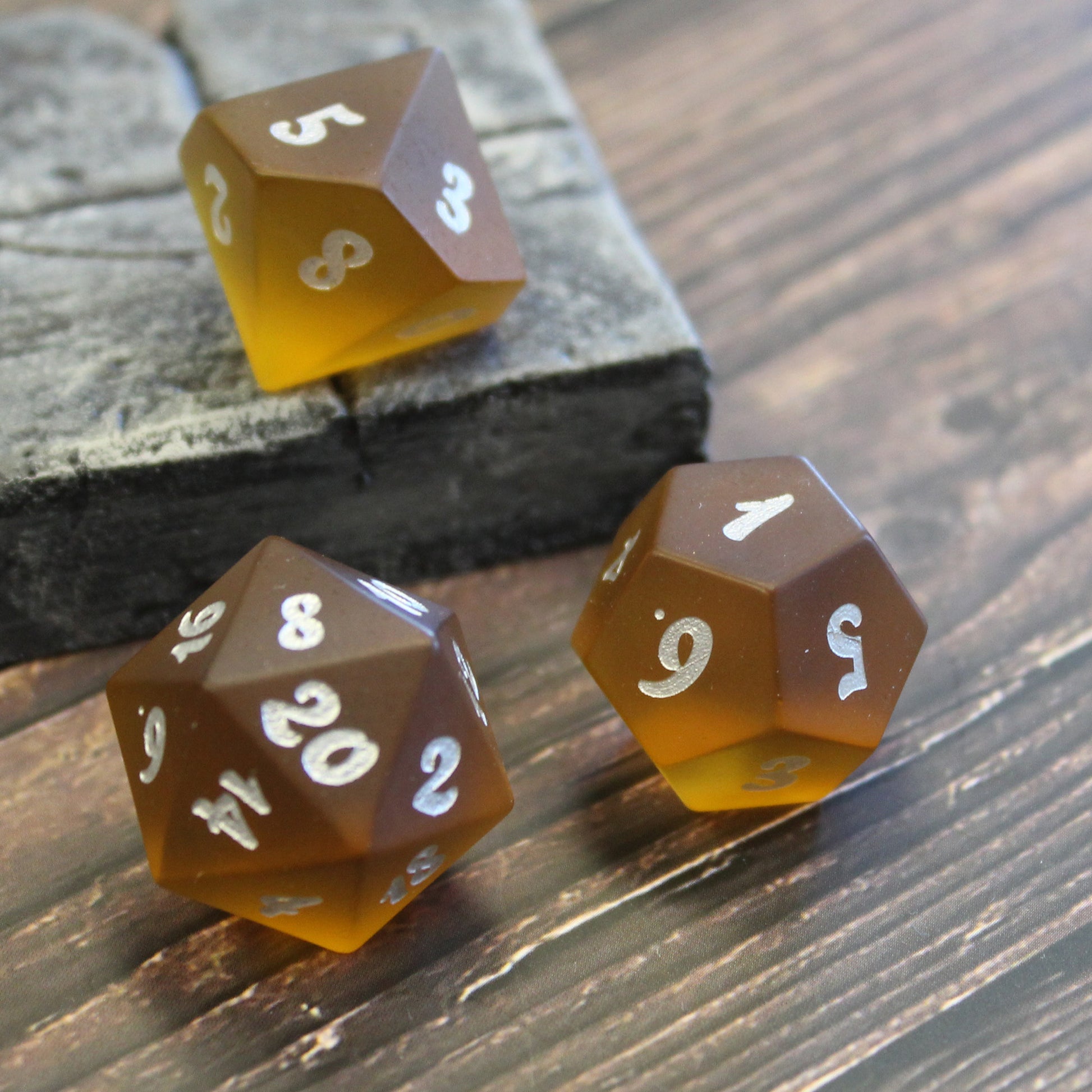 A few brown translucent glass RPG dice with silver numbering with light shining through them on a wooden surface.