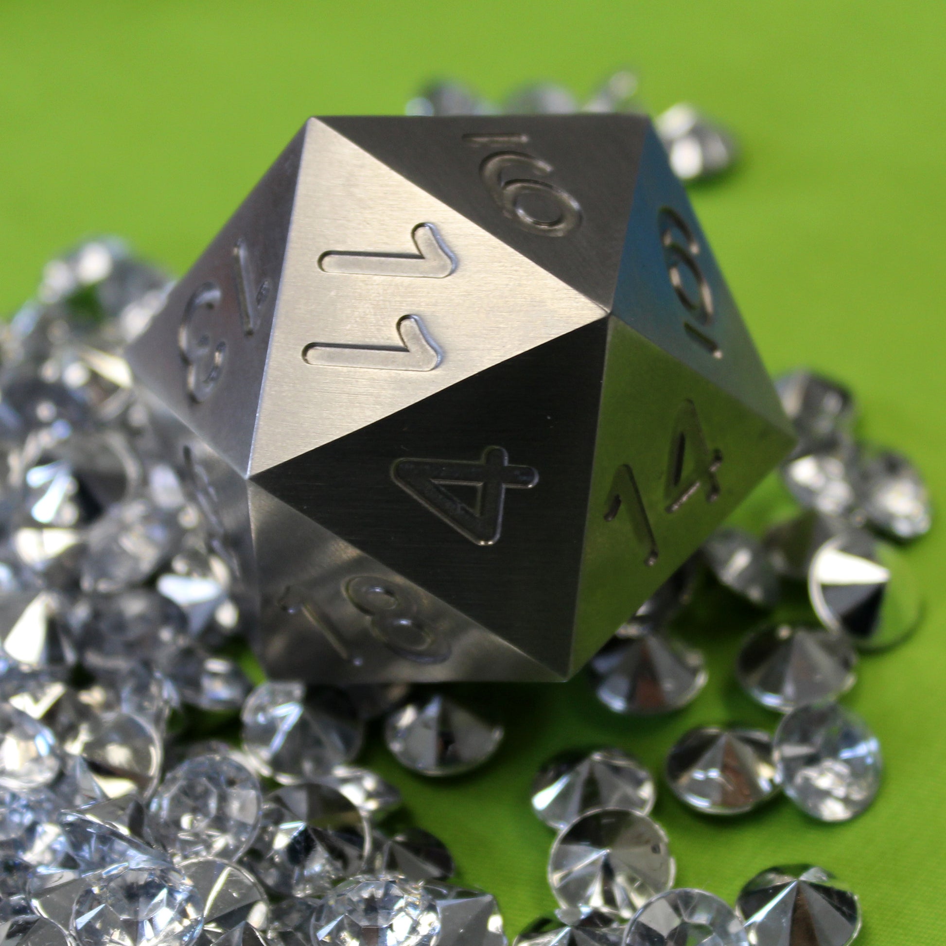 An oversized silver-colored metal twenty-sided die sitting on a pile of rhinestones on a green background.