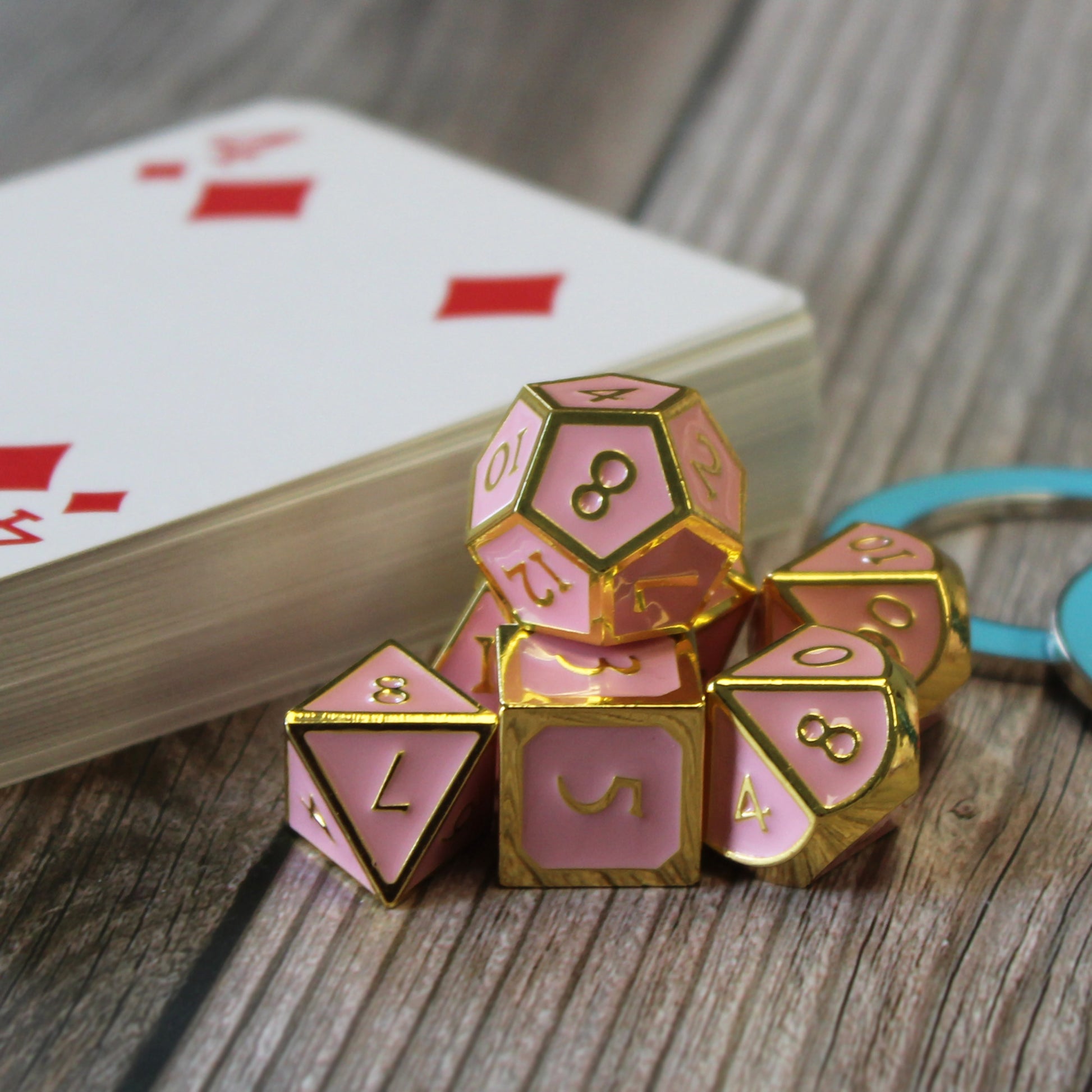 Metal Imperial Pink dice set arranged in a pile on a wooden surface, with the D12 on top. Behind the pile is a stack of playing cards with the 4 of diamonds on top, and two blue blank condition rings. 