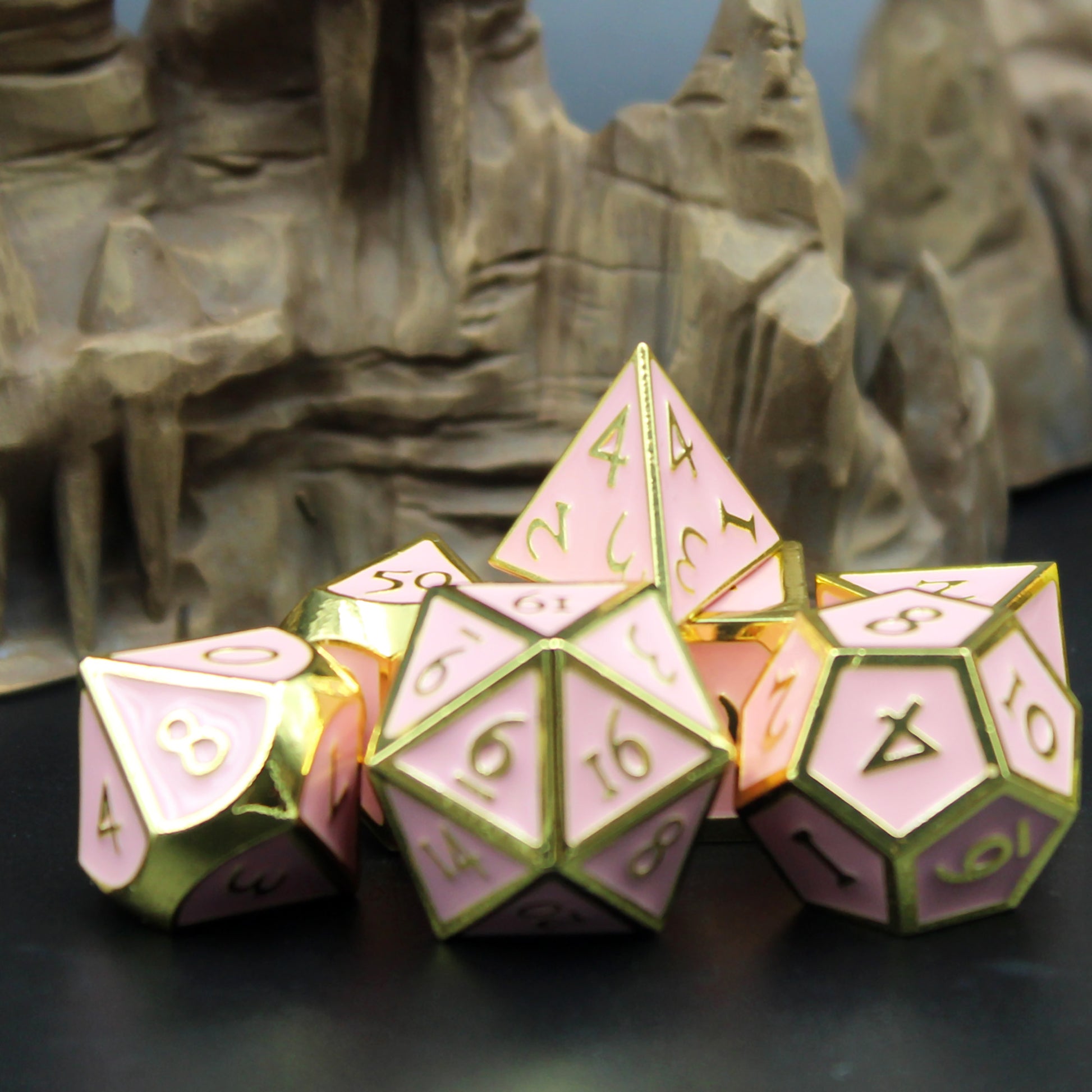 Metal Imperial Pink Dice set arranged in a pile on a black surface. The dice is set in front of a  rocky terrain set piece. 