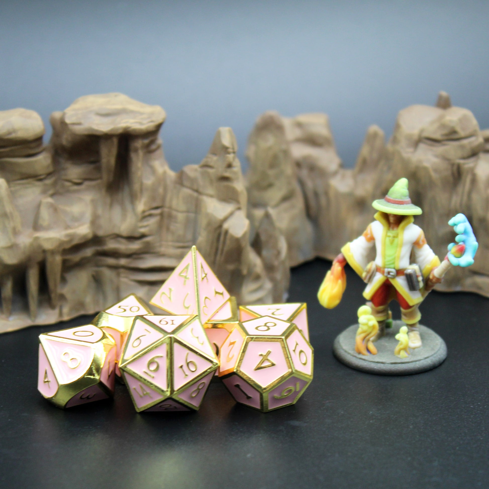 Metal Imperial Pink dice set arranged in a pile on a black surface next to a mini figure. Behind them is a rocky terrain set piece. 