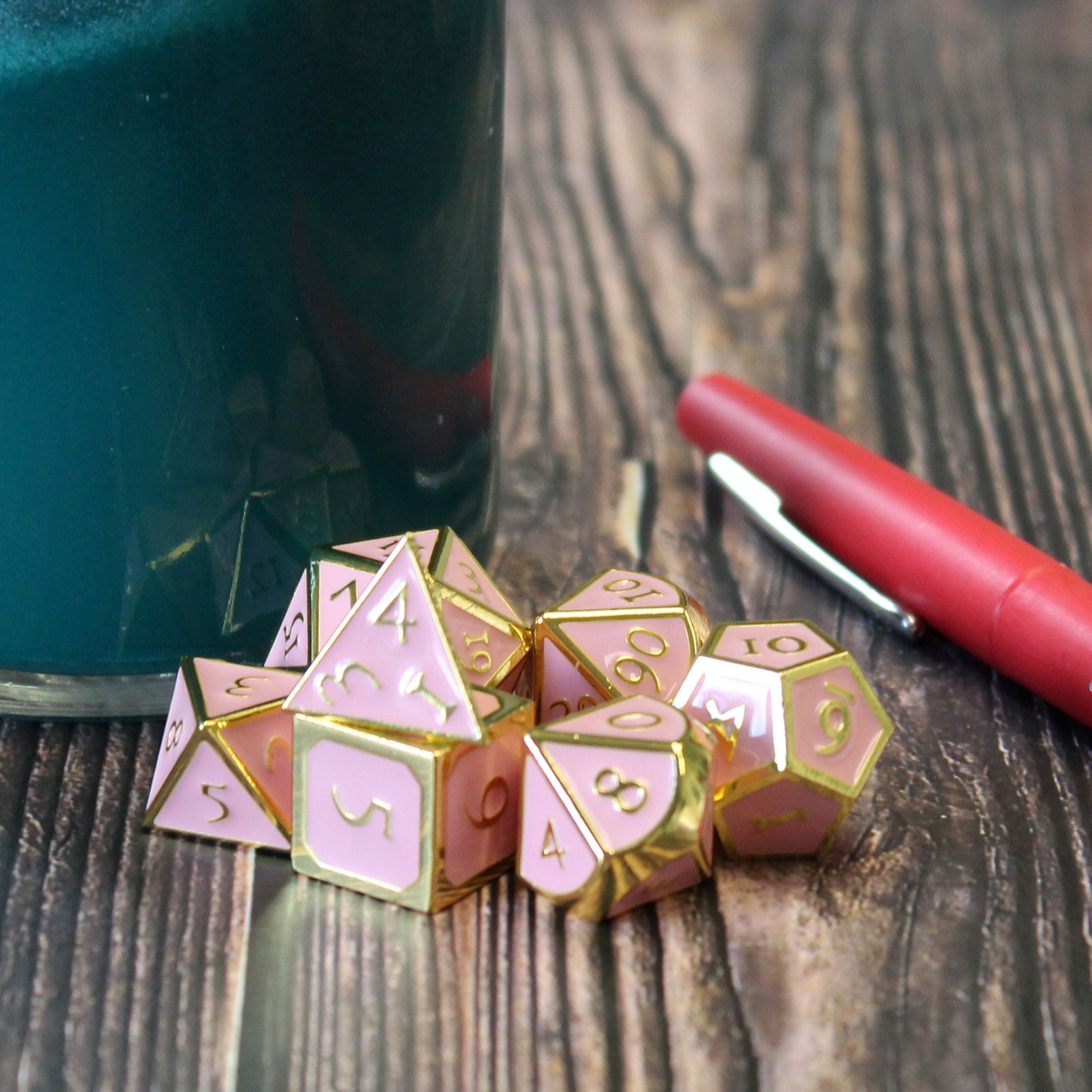 Metal Imperial Pink dice set arranged in a pile on a dark wood surface. Behind the pile is a blue glass on the left and a red pen on the right. 