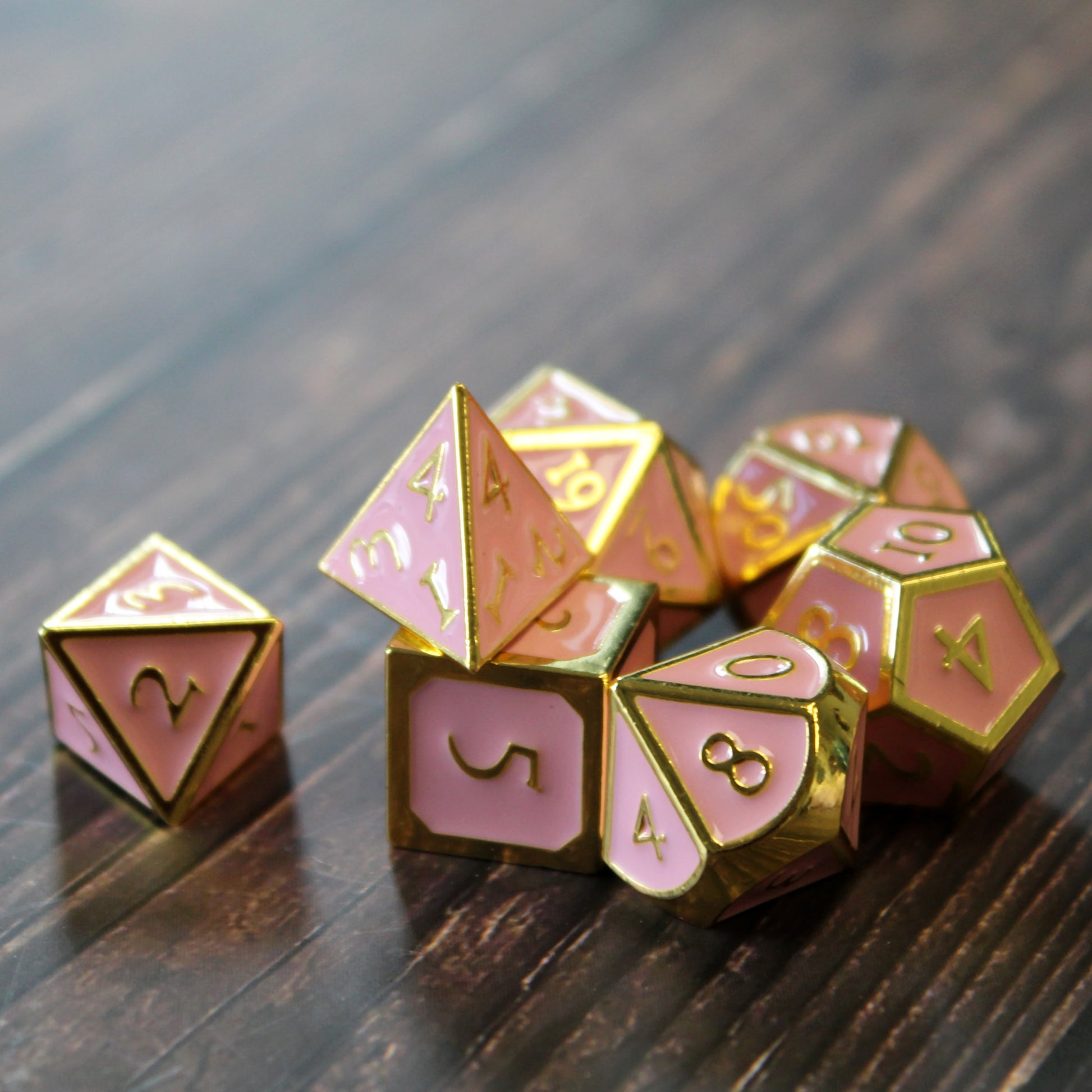 Metal Imperial Pink dice set arranged in a pile on a dark wood surface. The D4 has been placed on top of the other dice. 