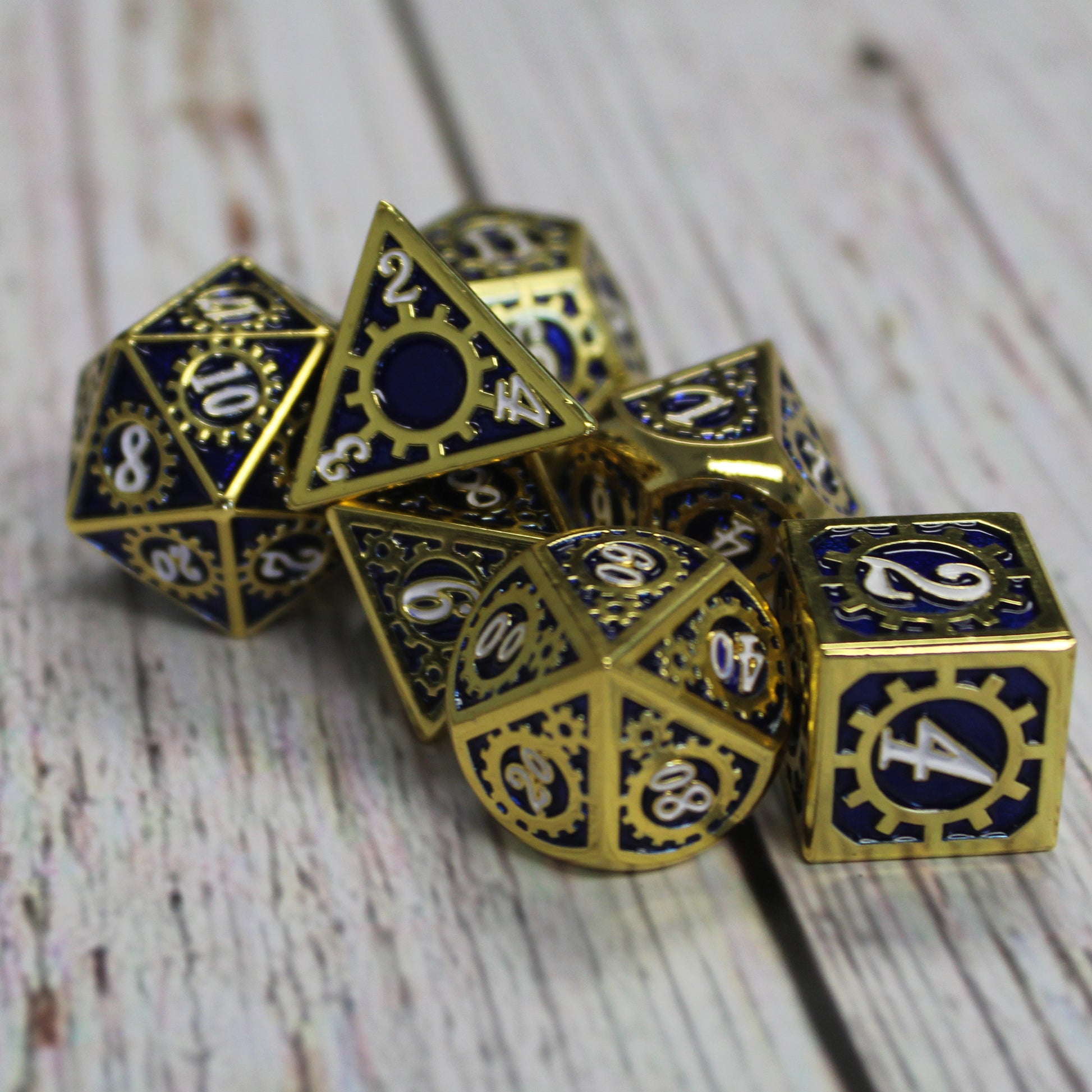 Mechanist blue and gold solid metal dice arranged in a pile on a wooden surface.  The d4 of this set rests atop with the numerals of the faces arranged around a cog.