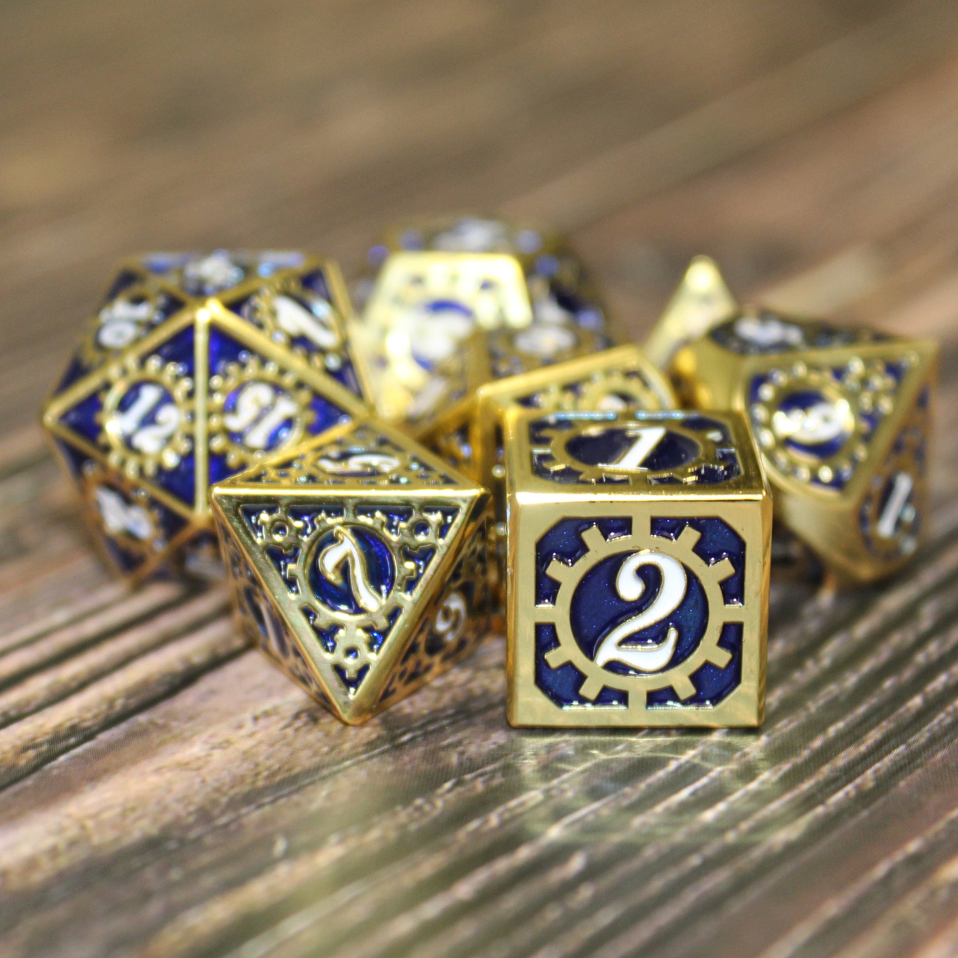 Metal Mechanist Blue and Gold dice set on a wooden surface with the d6 and d8 in the foreground.  The 2 face shows on the d6 with a white numeral ringed by a cog on a deep blue background framed by a raised golden edge.