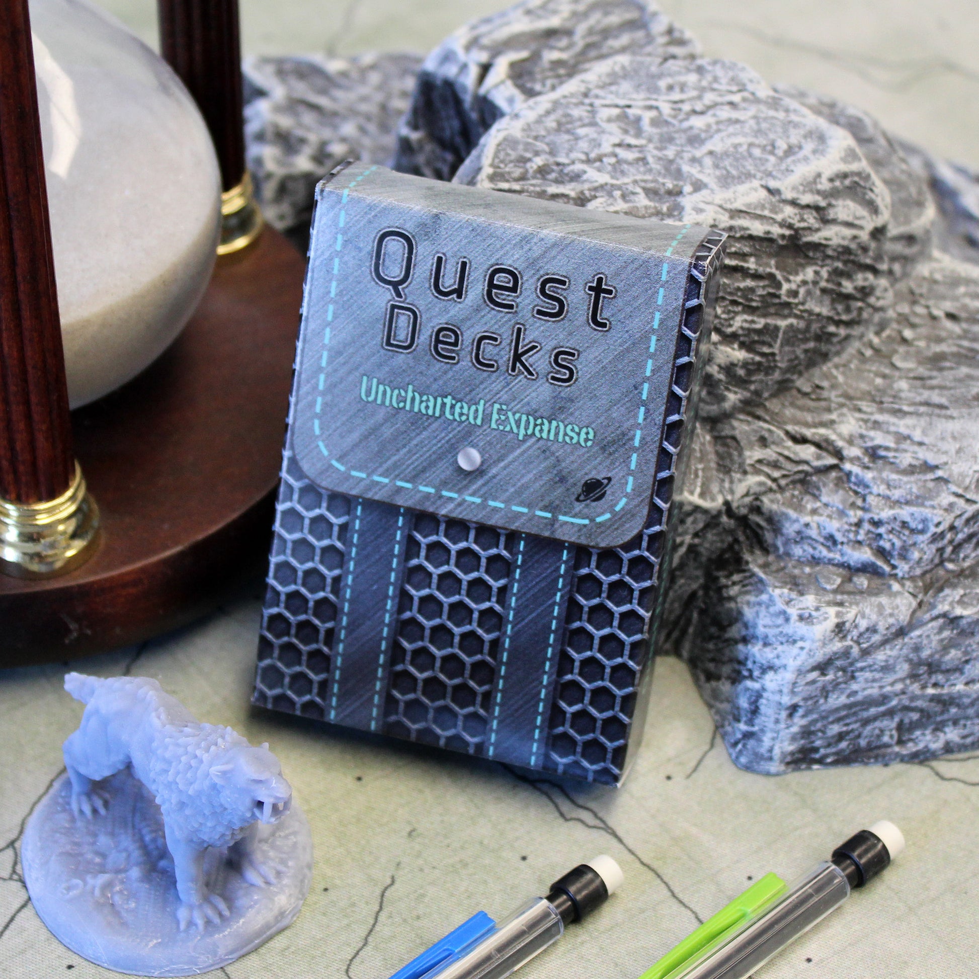 Picture of the Uncharted Expanse Quest Decks tuck box leaning against scatter terrain on a cloth map and a 2inch miniature.