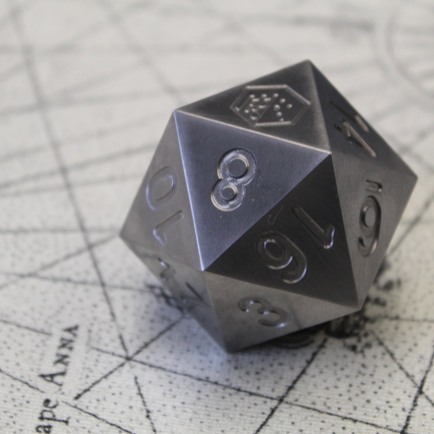 An oversized silver-colored metal twenty-sided die with a Dice Dungeons logo in place of the number twenty sitting on a fantasy map