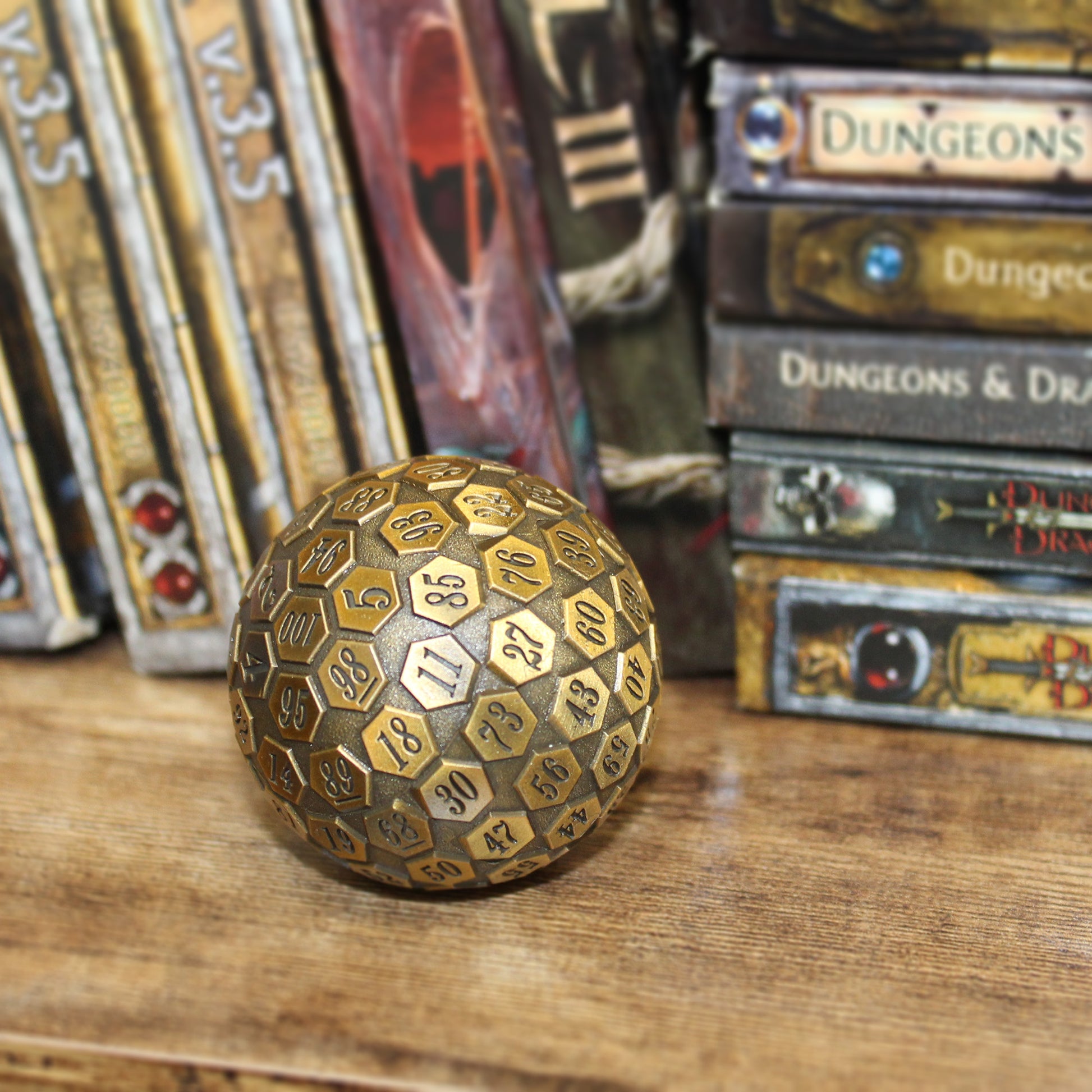 A 45mm diameter solid zinc alloy d100 with an antique gold finish resting on a shelf with various RPG sourcebooks in the background.