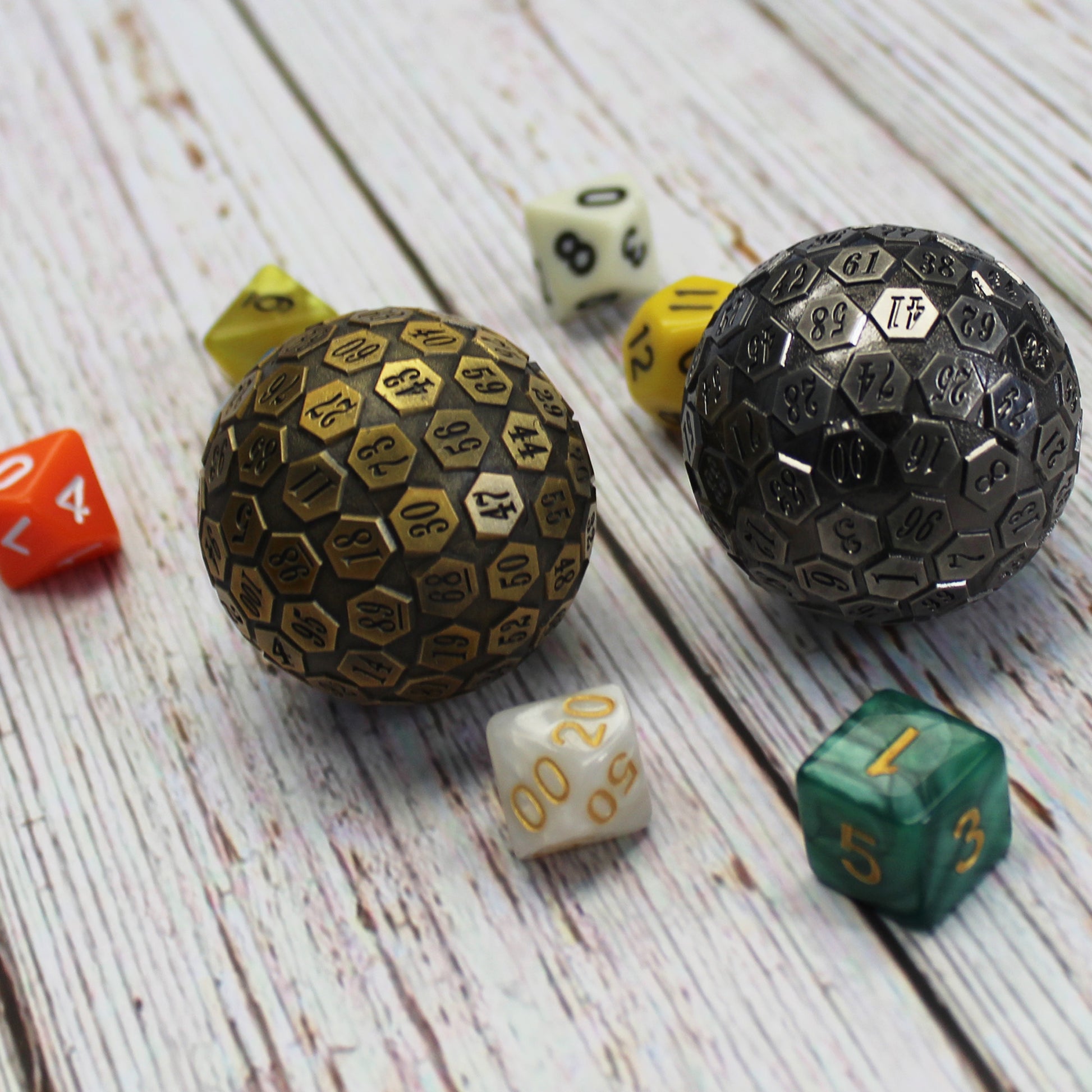 A comparison picture of the antique gold d100 beside the antique silver d100 resting on a wooden surface with standard sized dice beside each for scale.